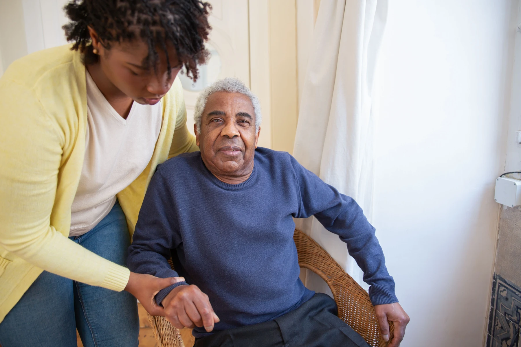 A caregiver beside a seated senior, offering assistance in a bright home setting